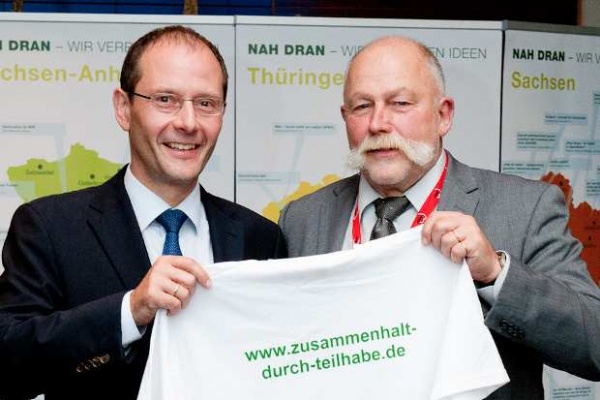 Der sächsische Innenminister Markus Ulbig und der AWO-Geschäftsführer Karl-Heinz Petersen freuen sich über den Einsatz der Demokratietrainer und halten ein weißes Demokratietrainer-T-Shirt hoch.